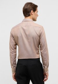 Camicia a maniche lunghe in tessuto marrone chiaro, con colletto classico, polsini abbottonati e superficie liscia; abbinata a pantaloni neri.