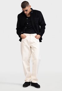 Camicia in corduroy nera, canotta nera aderente, pantaloni a gamba larga crema e scarpe slip-on nere. Sono visibili occhiali da sole e un dettaglio sulla tasca laterale.