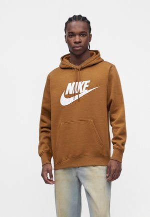 Jeune homme aux cheveux tressés portant un sweat à capuche Nike marron avec un logo blanc et un jean bleu clair, se tenant devant un fond blanc uni.