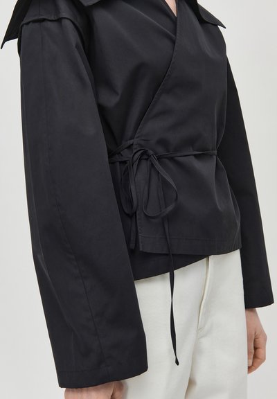 Black wrap jacket with long sleeves tied at the waist, paired with high-waisted white pants against a plain background.