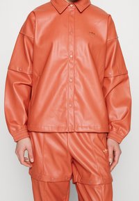 Giacca in similpelle arancione con collo, bottoni a pressione e polsini elasticizzati, abbinata a pantaloni coordinati. Branding discreto sulla giacca.