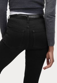 Schwarze Denim-Jeans mit enganliegendem Schnitt, ausgestattet mit zwei Gesäßtaschen und einem glatten schwarzen Lederbund. Der Stoff wirkt leicht strukturiert.