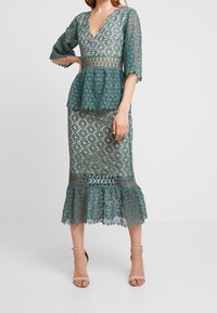 Teal spetsklänning med peplummidja, V-ringning och trekvartslånga ärmar, med en utsvängd fåll och intrikata cirkulära mönster.