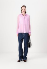 Hauchige, hellrosa Bluse mit Rüschensaum an Kragen und Ärmeln, kombiniert mit locker sitzenden dunkelblauen Jeans und einer kleinen schwarzen Handtasche. Sandalen runden das Outfit ab.