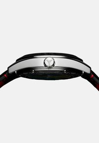 Silberfarbige Uhr mit schwarzem Gehäuse und griechischem Schlüsselmuster, transparentem Zifferblatt und roten Akzenten an einem schwarzen Band.