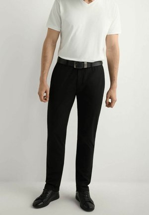 Mężczyzna ubrany w biały, krótki rękaw z dekoltem w serek, czarny pasek, wąskie czarne spodnie i czarne sznurowane buty, stojący na tle gładkiej ściany.