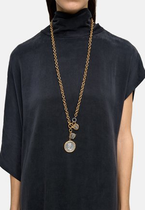 Długi złoty naszyjnik z trzema zawieszkami w kształcie monet, noszony na czarnej sukience z wysokim kołnierzem i krótkimi rękawami.