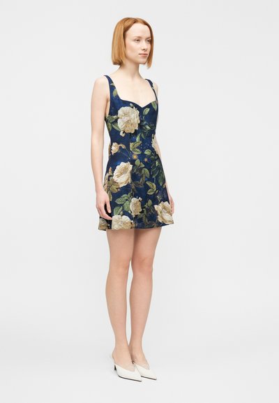 Jurk met bloemmotief op een marineblauwe achtergrond, skaterstijl, vierkante halslijn en dunne bandjes. Model draagt witte mules.