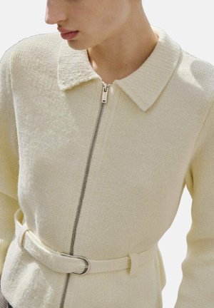 Chaqueta de punto color crema con cremallera, que cuenta con un cuello y una cintura ceñida con cinturón para dar forma. La textura es suave y ligeramente texturizada.