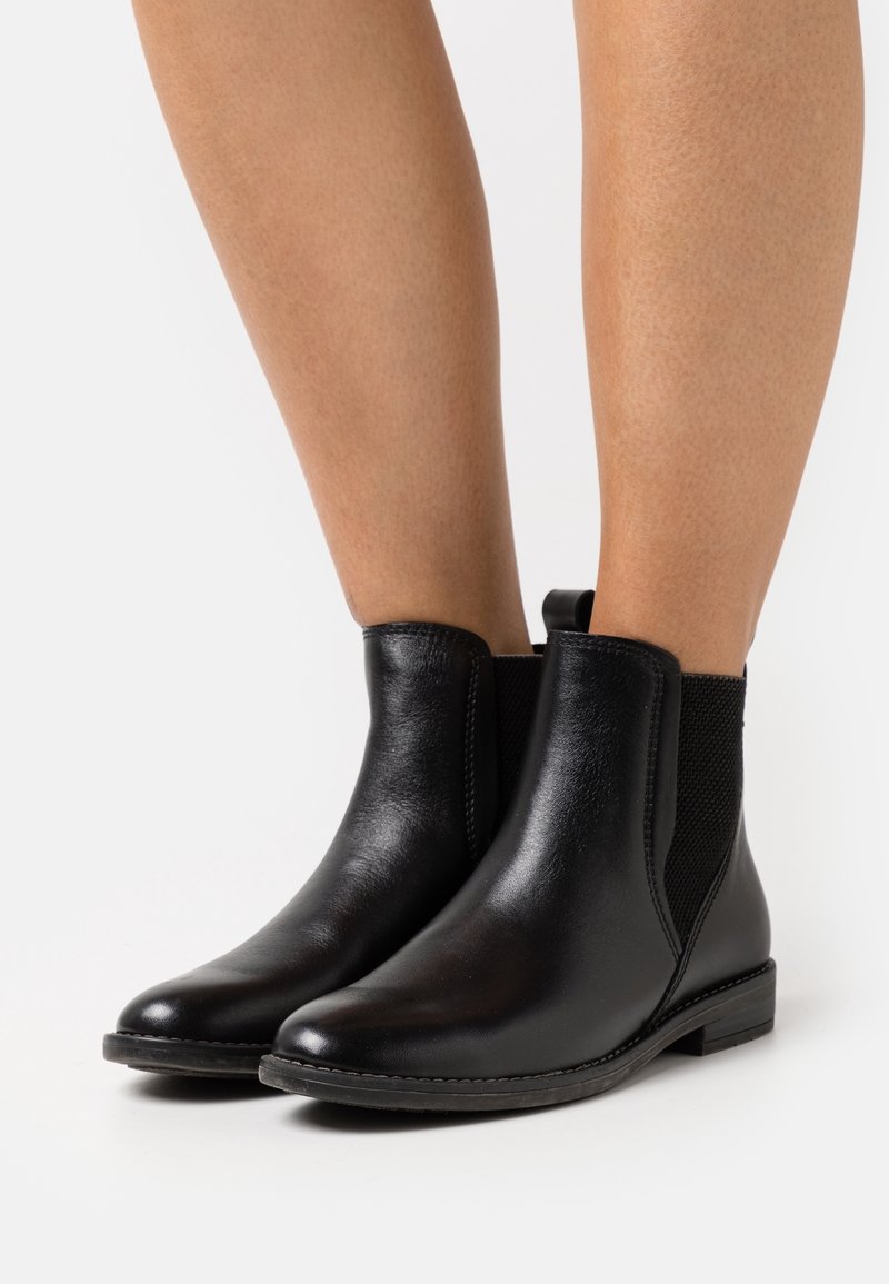 Bottes cheville en cuir noir avec panneaux latéraux élastiques portées sur jambes nues, présentées sur un fond clair uni.