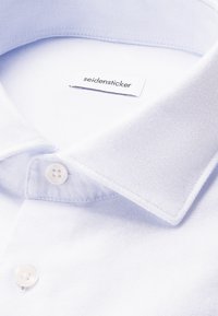 Lichtblauwe overhemdkraag van een textuurstof, met een genaaide label en een witte knop aan de onderkant. Eenvoudig ontwerp, zonder patronen.