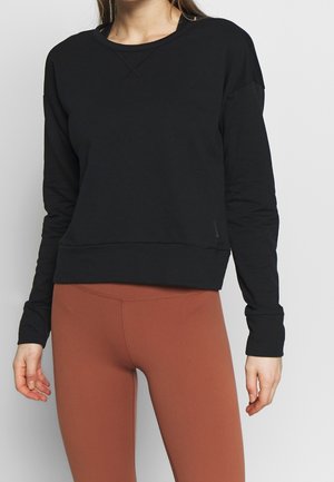 Kvinde iført en sort, langærmet cropped sweatshirt og rustfarvede, højttaljede leggings mod en neutral baggrund.