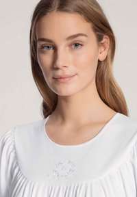 Blusa bianca con scollatura rotonda, maniche plissettate e ricamo floreale sul petto, realizzata in tessuto morbido con una texture liscia.