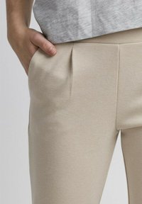 Beige op maat gemaakte broek met een textuurstof, voorzien van voorplooien en zijzakken. Een hand ligt in de zak.