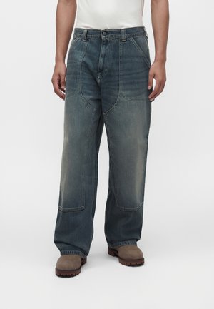 Uomo che indossa jeans blu sbiaditi e larghi con toppe rinforzate sulle ginocchia e stivali da lavoro marroni, in piedi davanti a uno sfondo semplice.