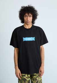 Camiseta de algodón negra con mangas cortas, que presenta un logo gráfico azul en el pecho. Combinada con pantalones de camuflaje.