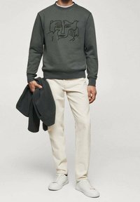 Mężczyzna w ciemnoszarej bluzie z abstrakcyjnym wzorem twarzy i ptaka, kremowe spodnie, białe trampki, trzymający w jednej ręce ciemnoszarą kurtkę.