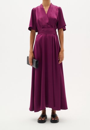 Femme portant une longue robe portefeuille violette à manches larges, tenant une pochette noire, et portant des sandales compensées noires, debout devant un fond blanc.