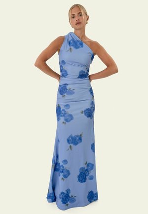 Frau trägt ein hellblaues, bodenlanges Kleid mit einem Schulterriemen und dunkelblauen Blumenmustern, steht mit Händen in der Hüfte.
