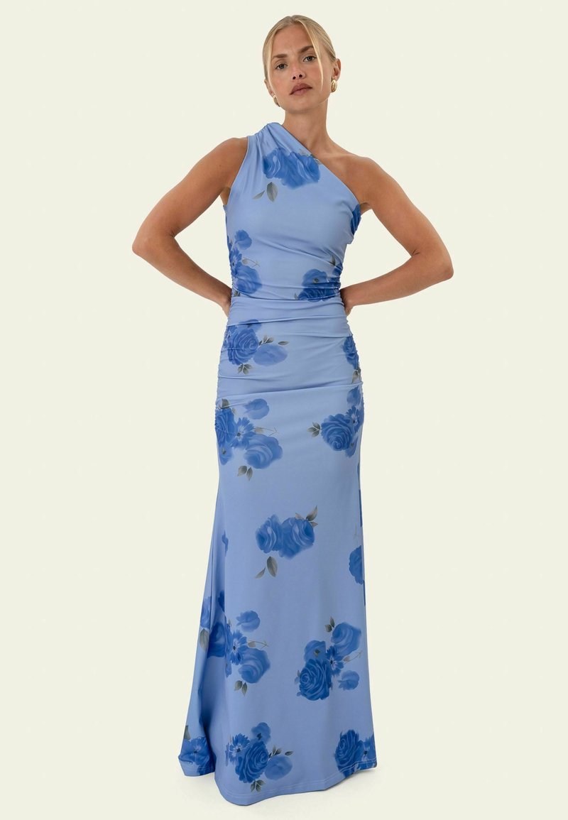 Femme portant une robe longue bleu clair à une épaule avec des motifs floraux bleu foncé, debout les mains sur les hanches.