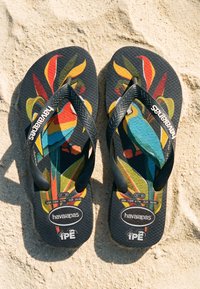 Paire de tongs noires Havaianas avec un motif perroquet coloré sur une surface sableuse, montrant les étiquettes de taille et les logos de la marque.