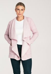 Jasnoróżowy kardigan o teksturze z otwartym przodem, długimi rękawami i dwiema kieszeniami, noszony na białej bluzce i zestawiony z ciemnymi dżinsami.