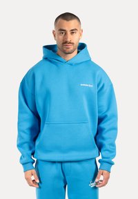 Hoodie bleu clair en tissu doux. Il présente une poche kangourou à l'avant et un logo blanc sur la poitrine. Coupe décontractée avec une capuche.