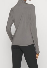 Šedé atletické tričko s dlouhým rukávem, vysokým límcem a úzkým střihem, vyrobené z hladké, lehké tkaniny. Prošívání podtrhuje strukturu.