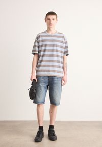 Stribet kortærmet t-shirt i lyseblå og brun, parret med denimshorts. Modellen holder en sort taske og har sorte sneakers og sokker på.