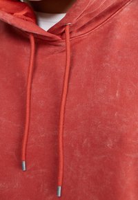 Sudadera con capucha roja hecha de tela suave con una textura ligeramente descolorida. Cuenta con dos cordones con terminales metálicos y un bolsillo frontal tipo canguro.