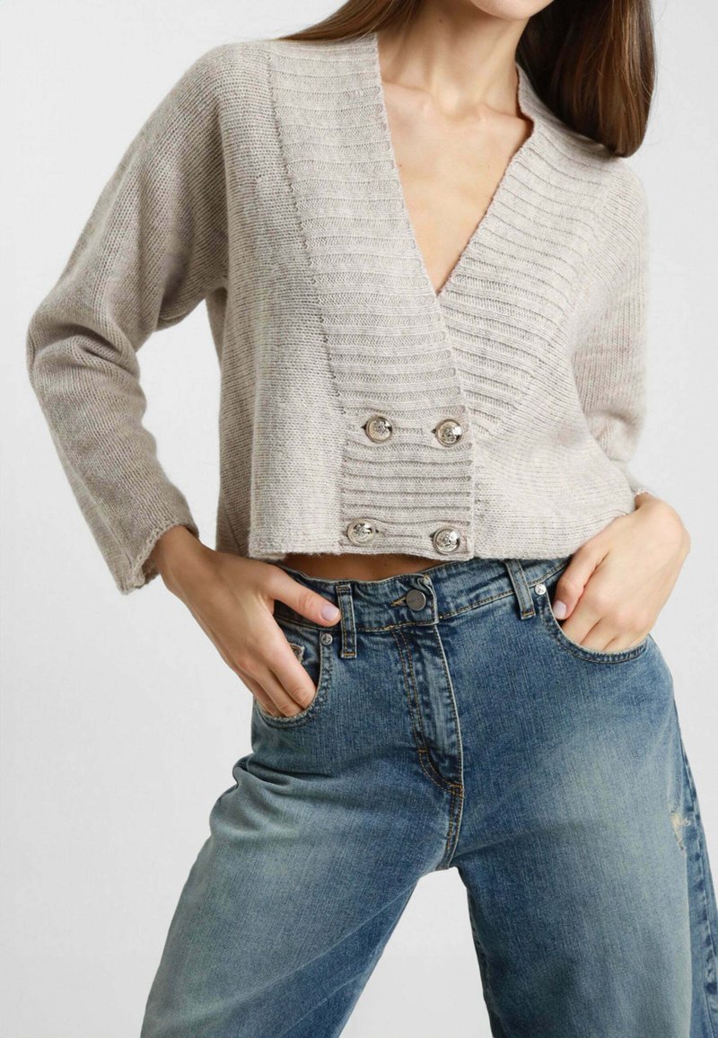 Maglione a maglia cropped beige con scollo a V profondo, texture a coste e doppi bottoni argentati, abbinato a jeans blu a vita alta.