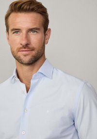 Lichtblauw katoenen shirt met button-down kraag en een klein wit embleem op de borst, met een gladde, gepolijste textuur.