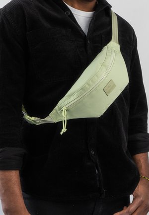 Borsa a tracolla verde chiaro con design triangolare, chiusura con cerniera, tracolla regolabile e dettaglio della superficie testurizzata. Toppa con logo sulla parte frontale.