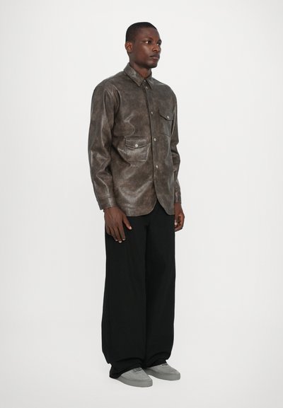 Chemise en cuir marron à boutons avec deux poches poitrine, surface texturée, associée à un pantalon large noir et des baskets grises.