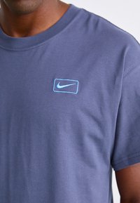 Nærbillede af en person iført en blå kortærmet skjorte med et lille lysblåt Nike swoosh-logo inden i et rektangel på brystet.