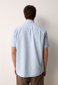 Camicia azzurro chiaro a maniche corte con righe bianche verticali, design con colletto, orlo dritto e tessuto morbido, indossata con pantaloni marroni.