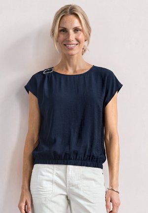 Femme blonde souriante, cheveux attachés en chignon bas, portant un haut sans manches bleu marine et un pantalon blanc devant un mur clair uni.