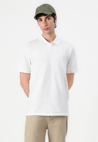 Weißes Baumwoll-Polohemd mit kurzen Ärmeln und einer Zwei-Knopf-Leiste, getragen mit hellbeigen Hosen und einer grünen Kappe. Schlichtes Design und glatte Textur.