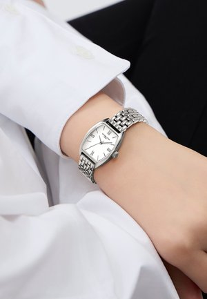 Montre en m�étal argenté avec un cadran rectangulaire courbé, un fond blanc, des chiffres romains et un bracelet en maillons texturés au poignet.