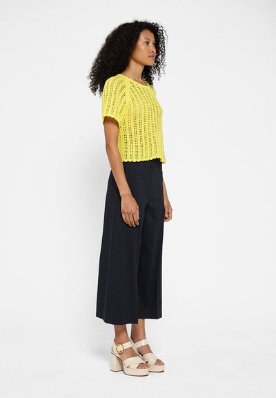 Top de punto amarillo de manga corta y escote redondeado, combinado con pantalones anchos negros y sandalias de tacón bloque en color beige.