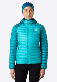 Kvinna klädd i en turkos uppblåsbar jacka med huva och matchande hårband, stående mot en enkel ljus bakgrund.