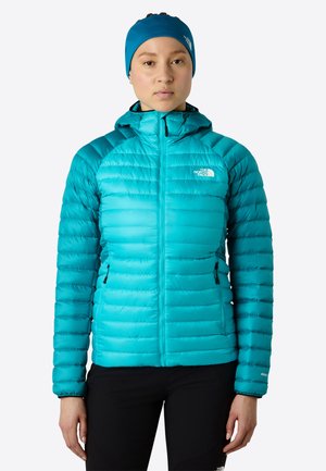 Vrouw met een teal pufferjack met capuchon en bijpassende hoofdband, staand tegen een effen lichte achtergrond.