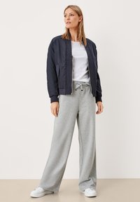 Vrouw staat rechtop in een marineblauwe bomberjack, witte T-shirt, grijze wijde sweatpants en witte sneakers tegen een effen achtergrond.