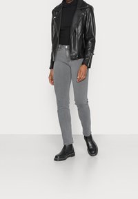 Mujer vestida con chaqueta de cuero negra de motociclista, suéter negro de cuello alto, jeans ajustados grises y botines negros de tobillo, de pie contra un fondo blanco.