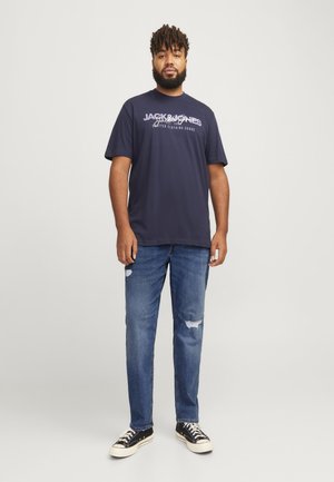 Jūras zilas kokvilnas t-krekls ar baltu grafisku tekstu, apvienots ar gaiši zili nolietoti džinsiem un melniem kanvas apaviem.