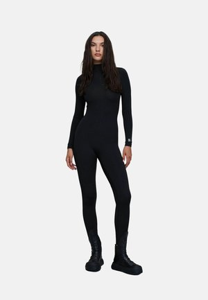 Femme aux longs cheveux foncés portant une combinaison noire cintrée à manches longues et des bottes à plateforme noires sur un fond clair uni.