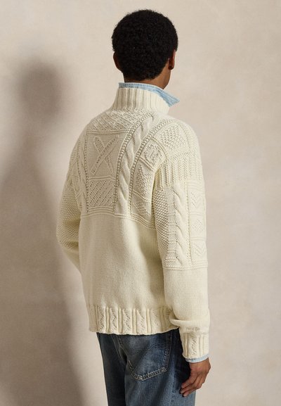 Άνδρας με σγουρά μαλλιά που φοράει εκρού πουλόβερ με πλεκτό σχέδιο και μπλε πουκάμισο με γιακά, όρθιος με την πλάτη σε απλό φόντο.