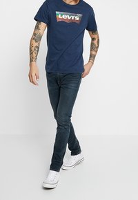 Mörkblå Levi's t-shirt med en färgglad logotypdesign, kombinerad med tajta mörka jeans och vita sneakers. Modellen har tatueringar på armarna.
