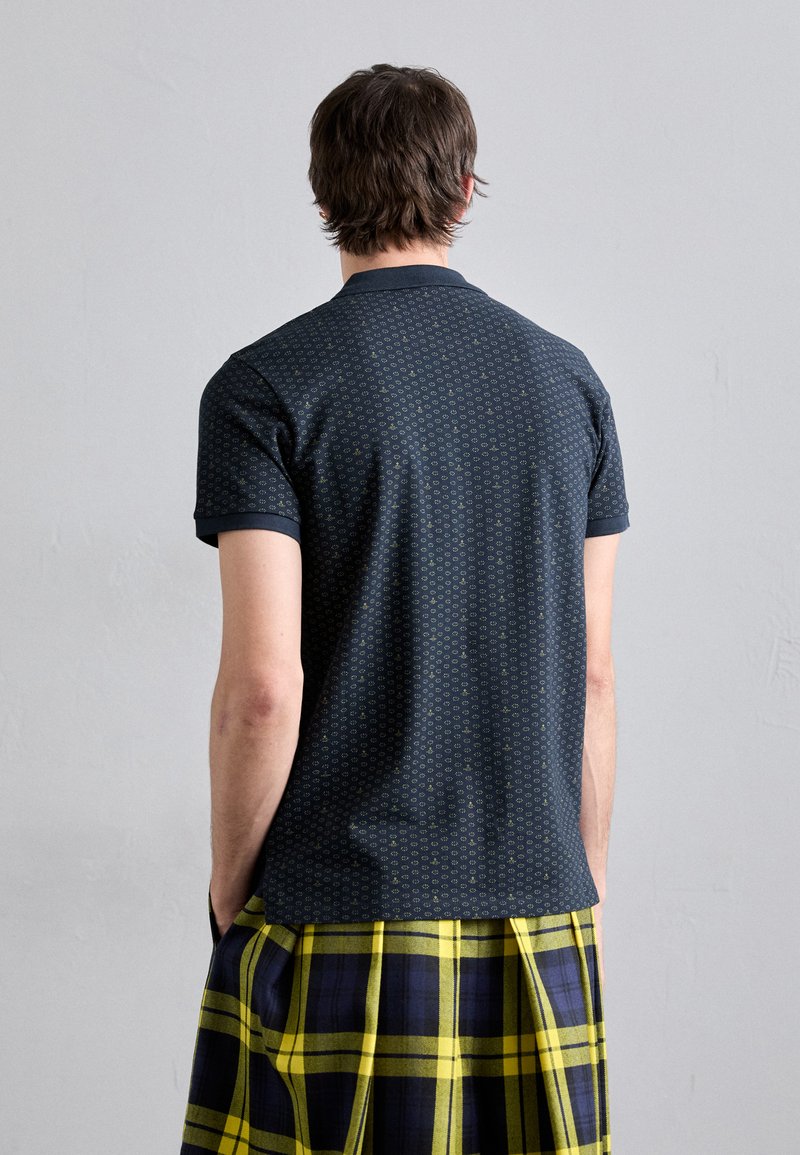 Marineblaues Polohemd mit einem strukturierten Muster aus kleinen Formen, kurzen Ärmeln und einem Kragen. Kombiniert mit einem karierten Rock in Gelb und Marineblau.
