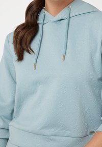 Felpa blu chiaro in tessuto strutturato con motivo logo in rilievo. Presenta un cappuccio con coulisse e dettagli in metallo color oro.